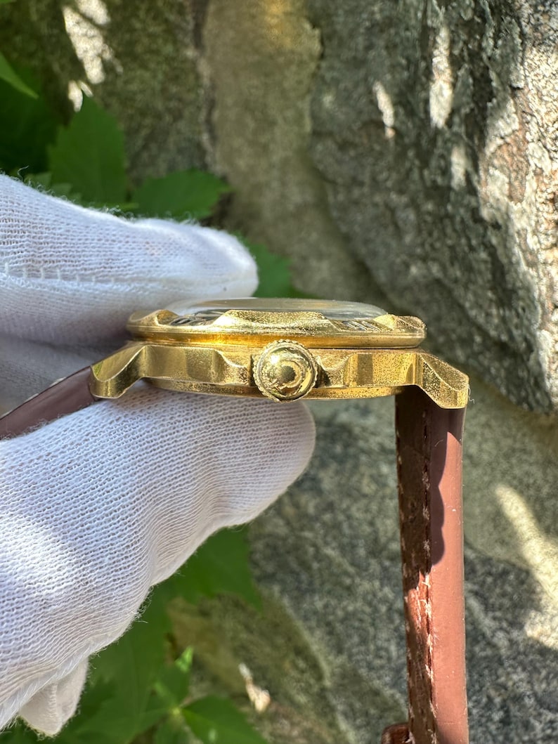 May include: Side view of a gold-toned wristwatch with a brown leather band. The watch has a textured face and a decorative crown. The watch is held by a gloved hand, showcasing the intricate details of the timepiece.