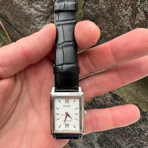 May include: A rectangular Orient watch with a white face, silver case, and black leather band. The watch face features black Roman numerals, a date window, and red hands. The band has a crocodile texture.