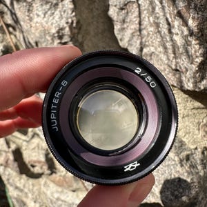 May include: A black Jupiter-8 2/50 camera lens with a silver ring and a small symbol. The lens has the text "JUPITER-8" and "2/50" printed on it. The lens is held in front of a blurred, rocky background.