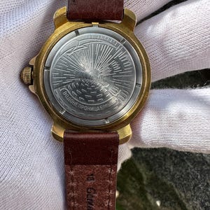 May include: Close-up of a vintage wristwatch with a gold-toned case and a brown leather strap. The watch face features a detailed design with a sunburst pattern. The strap is marked "18 Genuine Leather."