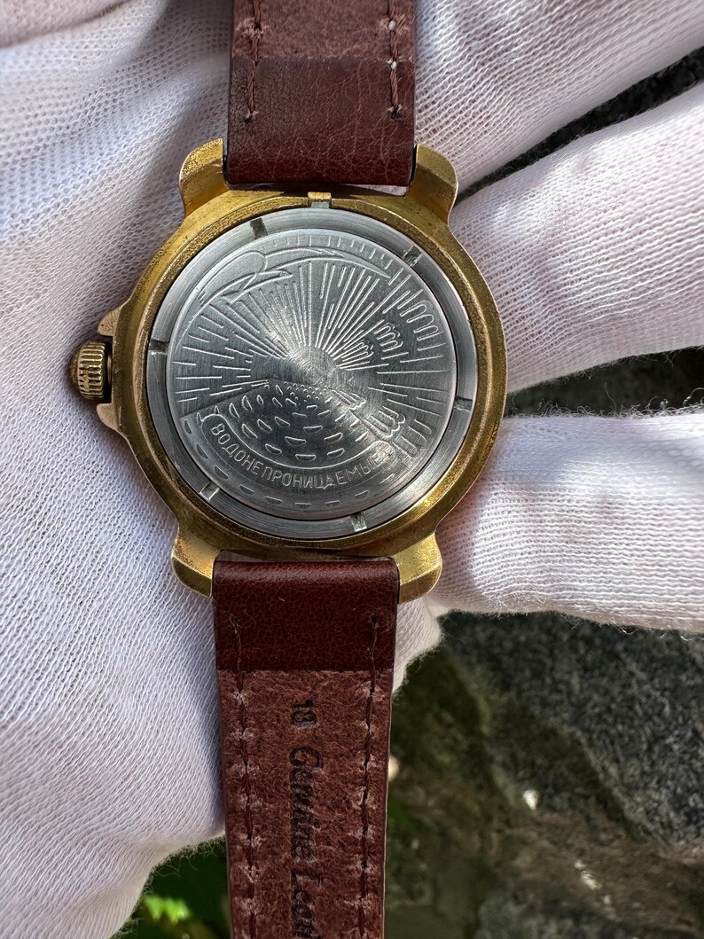 May include: Close-up of a vintage wristwatch with a gold-toned case and a brown leather strap. The watch face features a detailed design with a sunburst pattern. The strap is marked "18 Genuine Leather."