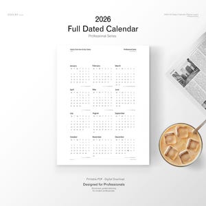 May include: A white 2026 full dated calendar with a professional series design. The calendar is open to display the months of the year. A glass of iced coffee and a newspaper are in the background.