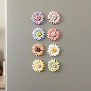 Peut inclure: Huit aimants en forme de marguerite aux couleurs pastel variées : lavande, rose, bleu, pêche, orange, blanc, jaune et vert. Chaque aimant a un centre texturé et est disposé sur une surface grise.