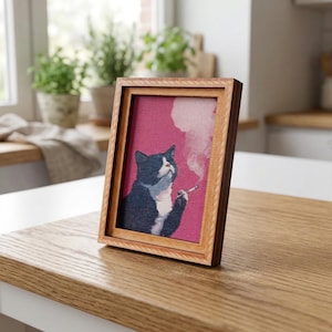 Peut inclure: Un cadre en bois clair contenant une image d'un chat noir et blanc fumant une cigarette sur fond rose. L'image est posée sur une table en bois clair, créant un effet de contraste.