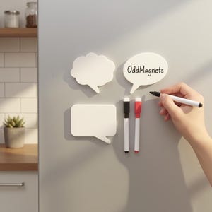 Op de afbeelding: Witte tekstballonmagneten met de tekst "OddMagnets" en twee markers, één zwart en één rood. Een hand houdt een zwarte marker vast. De magneten bevinden zich op een grijze ondergrond.