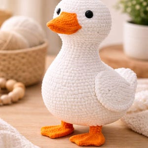 Könnte beinhalten: Eine handgefertigte weiße Häkelente mit orangefarbenem Schnabel und Füßen. Die Ente hat schwarze Augen und kleine Flügel. Das Plüschtier sitzt auf einer Holzoberfläche. Der Hintergrund ist verschwommen, mit Hinweisen auf Garn und eine Pflanze.