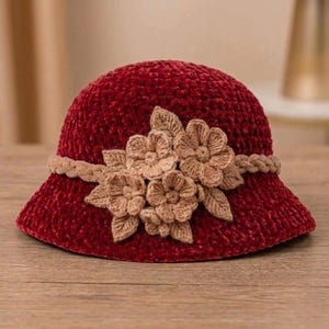 Puede incluir: Un sombrero cloche burdeos con un diseño floral de ganchillo. El sombrero tiene un ala ancha y una banda de adorno trenzado. Las flores son de color marrón claro y están unidas a la parte delantera del sombrero.