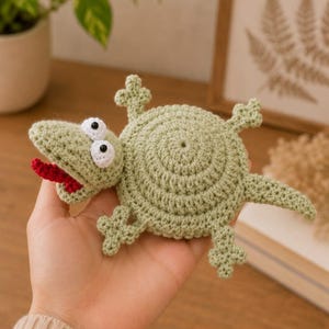May include: A light green crocheted lizard toy featuring a round body, four small legs with three-toed feet, a pointed tail, and a head with two white circular eyes containing black pupils and an open mouth showing a red crocheted tongue.