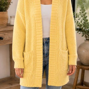 May include: A bright yellow open-front cardigan featuring a textured knit pattern, long sleeves with ribbed cuffs, a wide ribbed collar, and two large patch pockets on the front. The garment is worn over a white top and light blue denim jeans.