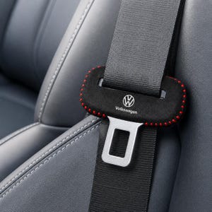 May include: A black seat belt cover featuring red stitched edges and the white Volkswagen logo with the text Volkswagen printed below it. The cover is positioned on a gray seat belt strap with a silver metal buckle visible below the cover.