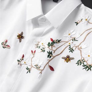 Può includere: Camicia bianca con ricami floreali e di insetti. Il design presenta fiori bianchi ricamati, foglie verdi, boccioli rossi, una farfalla e due api. La camicia ha un colletto classico e chiusura con bottoni.