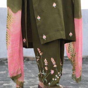Può includere: Pantaloni e top verde oliva ricamati con una sciarpa rosa e verde oliva. I pantaloni presentano ricami floreali lungo la gamba. Il top ha piccoli ricami floreali. La sciarpa ha un motivo rosa e verde oliva.