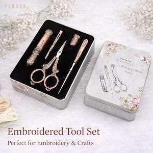 Könnte beinhalten: Ein Stickwerkzeug-Set in einer dekorativen Dose. Das Set enthält eine Schere, eine Nadel, einen Fingerhut und andere Werkzeuge mit aufwendigen Details im Vintage-Stil. Die Dose ist hellgrau mit floralen Akzenten.