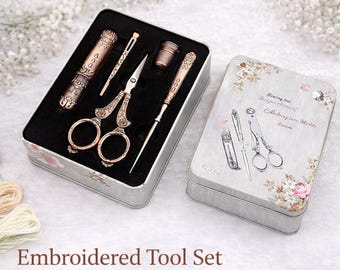 May include: An embroidered tool set in a decorative tin. The set includes scissors, a needle, a thimble, and other tools with ornate, vintage-style detailing. The tin is light gray with floral accents and the text "Embroidered Tool Set".