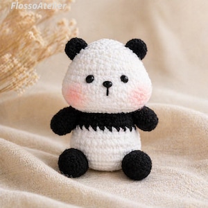 May include: A handmade crochet panda bear with a white body, black accents, and rosy cheeks. The panda has black button eyes and a small black nose. The arms and legs are also black. The panda is sitting upright.