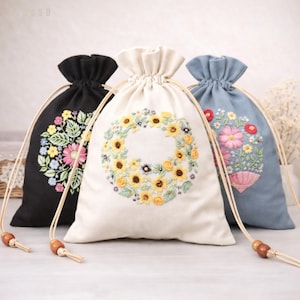 May include: Three drawstring bags with embroidered floral designs. One black bag features a colorful floral arrangement, a cream-colored bag has a sunflower wreath, and a blue bag showcases flowers in a pink vase. Each bag has a wooden bead detail.