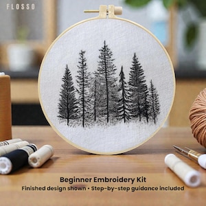 Può includere: Un telaio da ricamo con una scena di foresta ricamata in bianco e nero. Il testo "Beginner Embroidery Kit" è visibile, insieme a "Finished design shown • Step-by-step guidance included". Sono visibili anche fili e aghi da ricamo.