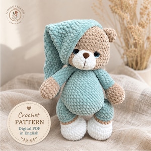 Peut inclure: Un ours en peluche crocheté en fil texturé doux dans des tons beige, bleu clair et blanc. L'ours porte un bonnet de nuit et un pyjama bleu clair assortis avec des pieds blancs. Une étiquette circulaire affiche le texte Crochet PATTERN Digital PDF in English.
