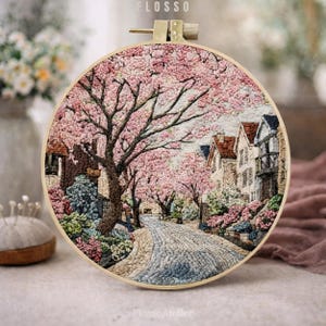 Può includere: Un telaio da ricamo dettagliato con una scena di strada con alberi di fiori di ciliegio in piena fioritura. L'opera raffigura una strada acciottolata fiancheggiata da case e alberi di fiori rosa. Il telaio è in legno e ha la parola "FLOSSO" in alto.