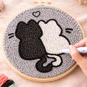 以下が含まれることがあります： グレーの丸いパンチニードル刺繍枠。黒と白の2匹の猫のシルエットと、その上にハートが描かれています。猫は灰色の背景に配置され、枠は木製です。手はパンチニードルツールを持っています。