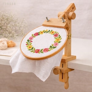 May include: A wooden embroidery hoop stand holds a white fabric with a floral cross-stitch design. The cross-stitch features a wreath of pink, yellow, and green flowers. The stand is made of light-colored wood and has adjustable features.