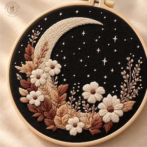 May include: A circular embroidery hoop containing black fabric with a crescent moon design stitched in cream thread. The moon is decorated with cream and brown floral motifs, including leaves and flowers, alongside small stitched stars in cream.
