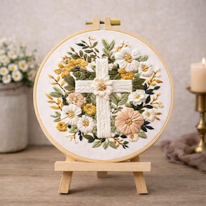 Könnte beinhalten: Gesticktes Kreuz, umgeben von Blumen in einem Holzreifen. Das Kreuz ist weiß, mit einem Blumendesign in der Mitte. Die Blumen sind weiß, gelb und pfirsichfarben, mit grünen Blättern. Die Stickerei wird auf einer kleinen Holzstaffelei präsentiert.