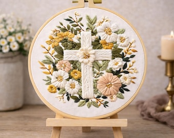 May include: Embroidered cross surrounded by flowers in a wooden hoop. The cross is white, with a floral design at the center. The flowers are white, yellow, and peach, with green leaves. The embroidery is displayed on a small wooden easel.
