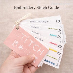 Può includere: Una mano tiene in mano una guida ai punti per ricamo. La guida presenta una scheda rosa con la parola "STITCH" e altri elementi decorativi. Mostra vari esempi di punti con nomi come "Punto indietro" e "Punto catenella", insieme a numeri.