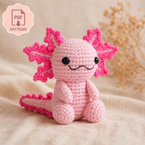 Peut inclure: Un jouet axolotl rose au crochet fait à la main, avec des branchies et une queue rose vif. L'axolotl a des yeux de sécurité noirs et une bouche cousue. Une icône de modèle PDF est dans le coin supérieur gauche.