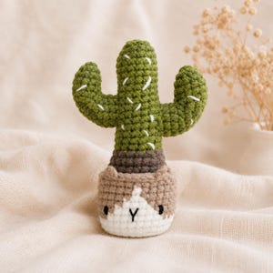 Puede incluir: Un juguete de cactus tejido a ganchillo con un cuerpo verde y detalles cosidos en blanco, colocado en una maceta marrón y blanca. La maceta tiene una pequeña cara bordada con dos ojos negros y una boca en forma de Y, hecha con puntos texturizados.