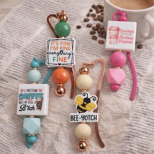Può includere: Diversi segnalibri decorativi con perline colorate e dettagli metallici. I segnalibri presentano varie frasi, tra cui "It's Fine, I'm Fine, Everything's Fine" e "Bee-Yotch". Uno ha un design a tazza di caffè.