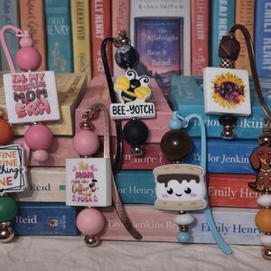 May include: A collection of decorative bookmarks with various designs and text. The bookmarks feature colorful beads, charms, and phrases like "It's Fine I'm Fine Everything's Fine" and "Bee-Yotch". They are displayed in front of a backdrop of books.