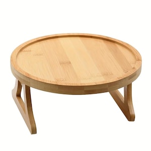 May include: A round, light-colored bamboo serving tray with folding legs. The tray has a raised edge and is supported by two V-shaped legs. The natural wood grain is visible throughout the tray's surface.