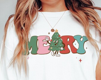 May include: White t-shirt with a colorful "Merry" graphic featuring a cartoon Christmas tree with a smiling face and waving hand.