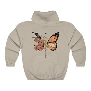 May include: Beige hoodie featuring a butterfly design with orange and black wings, floral accents, and the text "there is magic in all of us." The design includes a heart and a splatter effect.