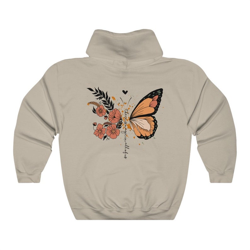 May include: Beige hoodie featuring a butterfly design with orange and black wings, floral accents, and the text "there is magic in all of us." The design includes a heart and a splatter effect.
