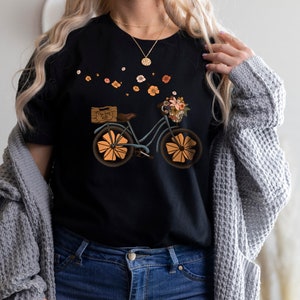 May include: Black t-shirt featuring a whimsical bicycle design. The bicycle is light blue with orange wheels and a basket filled with flowers. The text on the wooden box reads "Free Your Mind". A floral arrangement is in the basket.