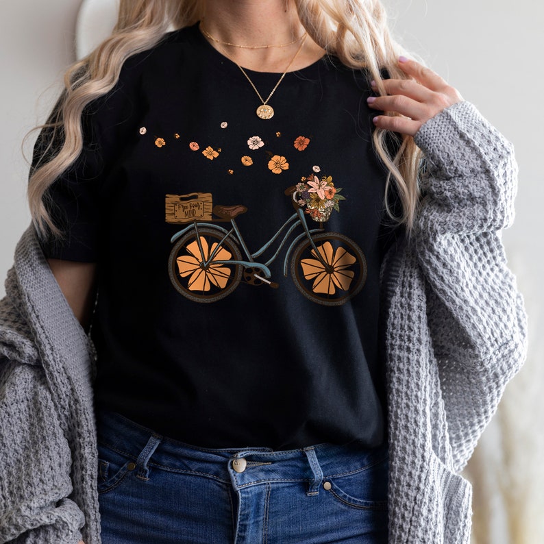 May include: Black t-shirt featuring a whimsical bicycle design. The bicycle is light blue with orange wheels and a basket filled with flowers. The text on the wooden box reads "Free Your Mind". A floral arrangement is in the basket.