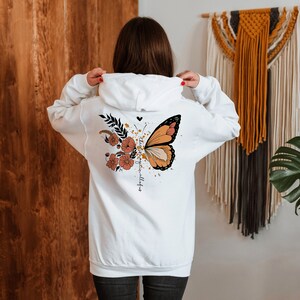 May include: White hoodie with a butterfly and floral design on the back. The butterfly is orange, black, and cream, with a floral arrangement in shades of orange and brown. The text "the best is yet to come" is printed on the back.