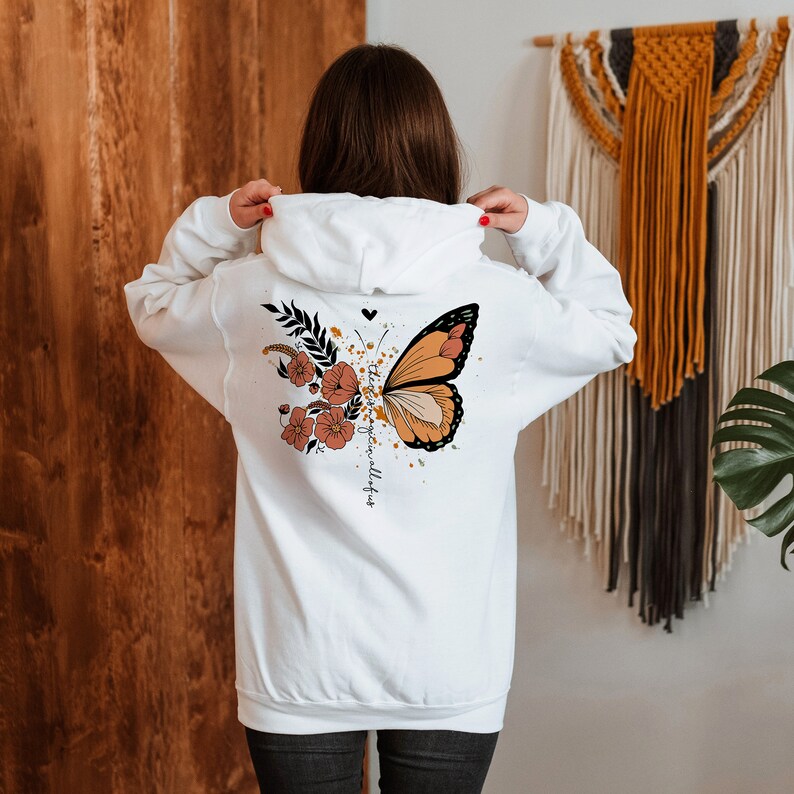 May include: White hoodie with a butterfly and floral design on the back. The butterfly is orange, black, and cream, with a floral arrangement in shades of orange and brown. The text "the best is yet to come" is printed on the back.