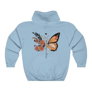 May include: Light blue hoodie featuring a butterfly design. The butterfly is orange and black, with floral and leaf accents. The text "there is magic in all of us" is written vertically on the right side of the butterfly. The hoodie has a drawstring hood.