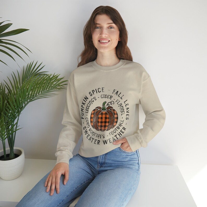 May include: A beige sweatshirt with a graphic of a pumpkin with a black and orange plaid pattern. The graphic is surrounded by text that reads "Pumpkin Spice - Cider - Leggings - Corn Bonfires - Sweater Weather - Fall Leaves - Flannel - Football".