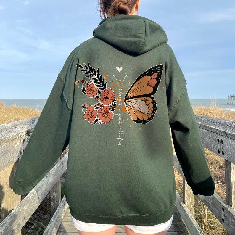 May include: Dark green hoodie with a butterfly and floral design on the back. The butterfly wing is brown, tan, and black. The floral design includes orange and white flowers. The text "there is magic in all of us" is printed vertically.