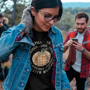 May include: A black sweatshirt with a graphic of a pumpkin with a plaid pattern. The text around the pumpkin reads "Spice - Cider - Flannel - Leggings - Bonfires - Football - Weather".