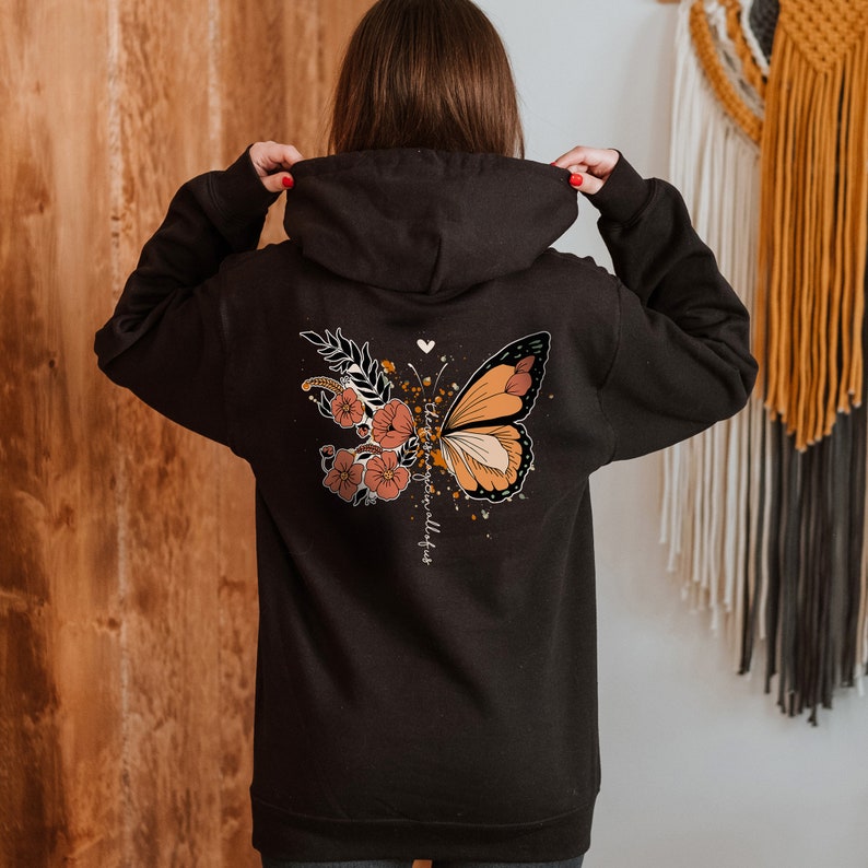 May include: Black hoodie featuring a butterfly and floral design on the back. The butterfly has orange, brown, and white details. The design includes the text "the magic is all of us".