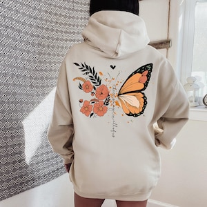 May include: Cream-colored hoodie featuring a butterfly design with orange and black wings, flowers, and the text "there is magic in all of us." The design includes a heart and floral elements.