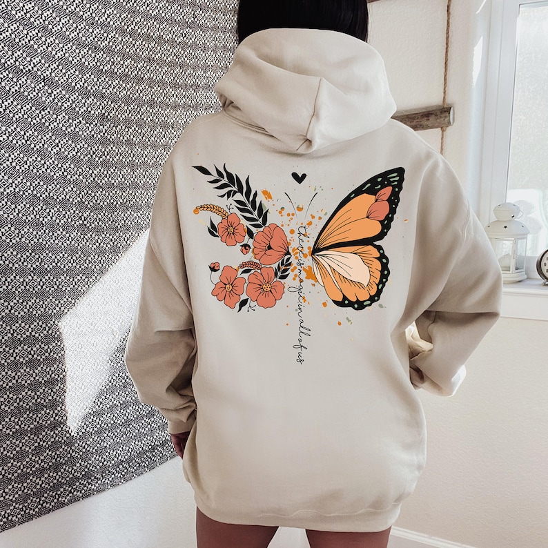 May include: Cream-colored hoodie featuring a butterfly design with orange and black wings, flowers, and the text "there is magic in all of us." The design includes a heart and floral elements.