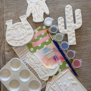 Pode incluir: Um kit de pintura DIY com figuras de cerâmica branca, incluindo uma ferradura, cacto, bota de cowboy, cartas de jogar e uma pessoa com chapéu. Inclui uma paleta de plástico branca, dois pincéis azuis e uma tira com seis potes de tinta.
