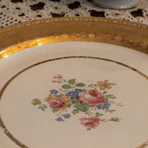 May include: A round, vintage plate with a gold rim and a white center. The center features a floral design with pink, blue, and yellow flowers. The plate is likely made of ceramic or porcelain.